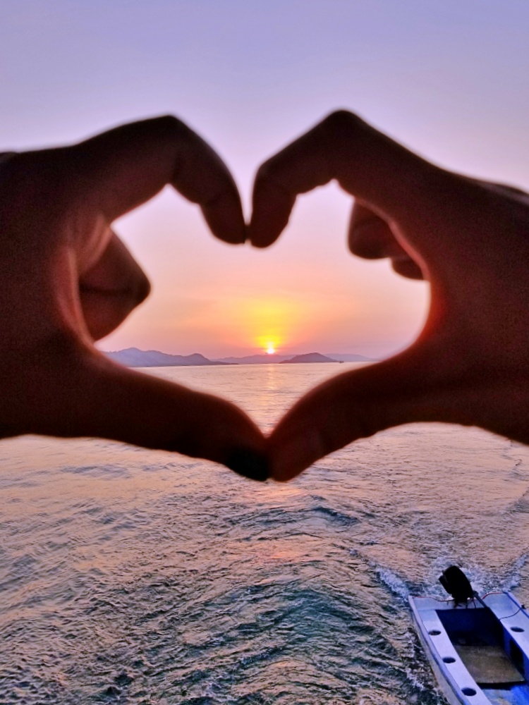 fingers making a heart shape with sun setting in the middle whilst on Komodo trip on boat